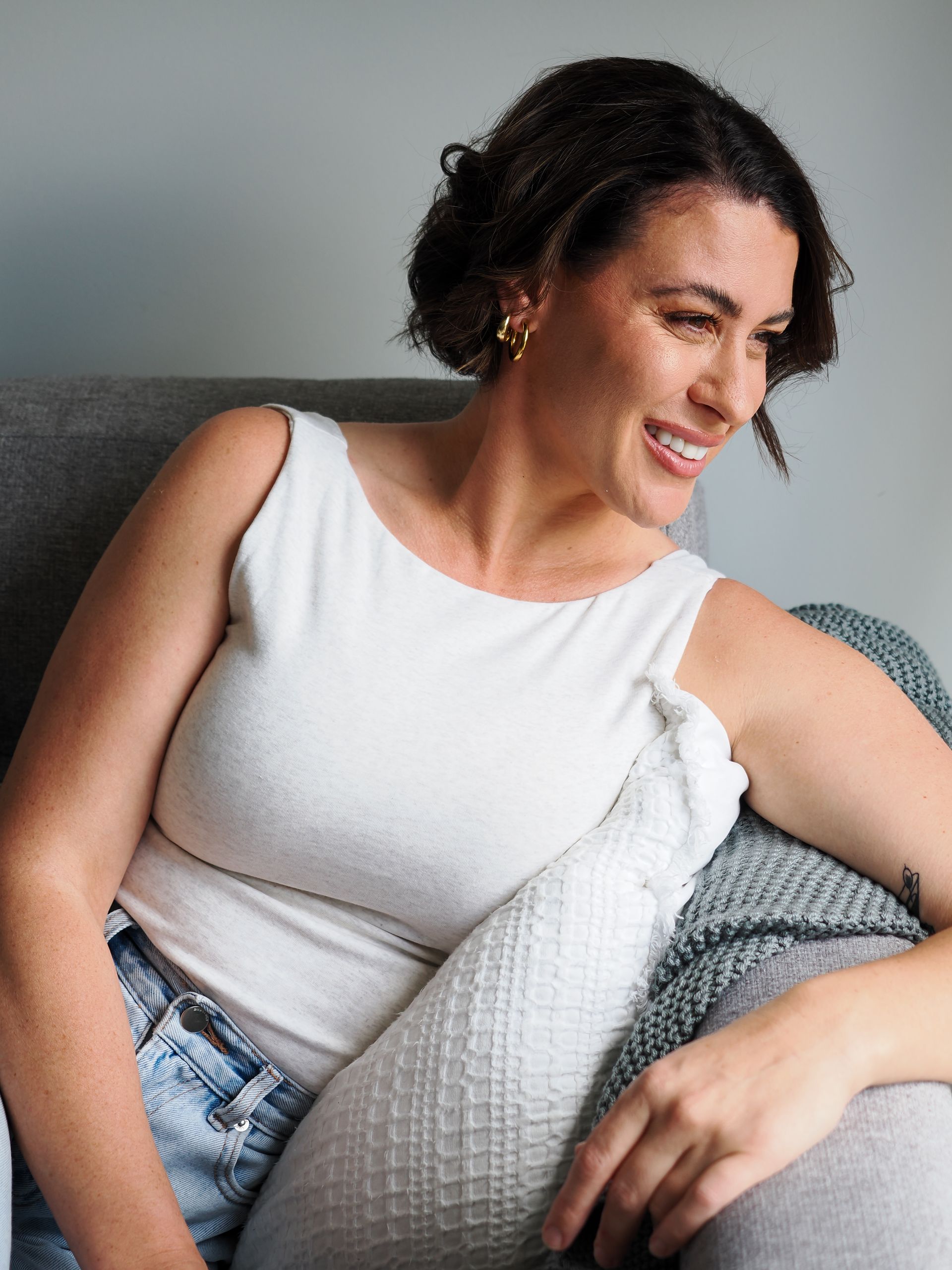 Woman with short dark hair smiles, sitting on a gray couch, wearing a white tank top and jeans.