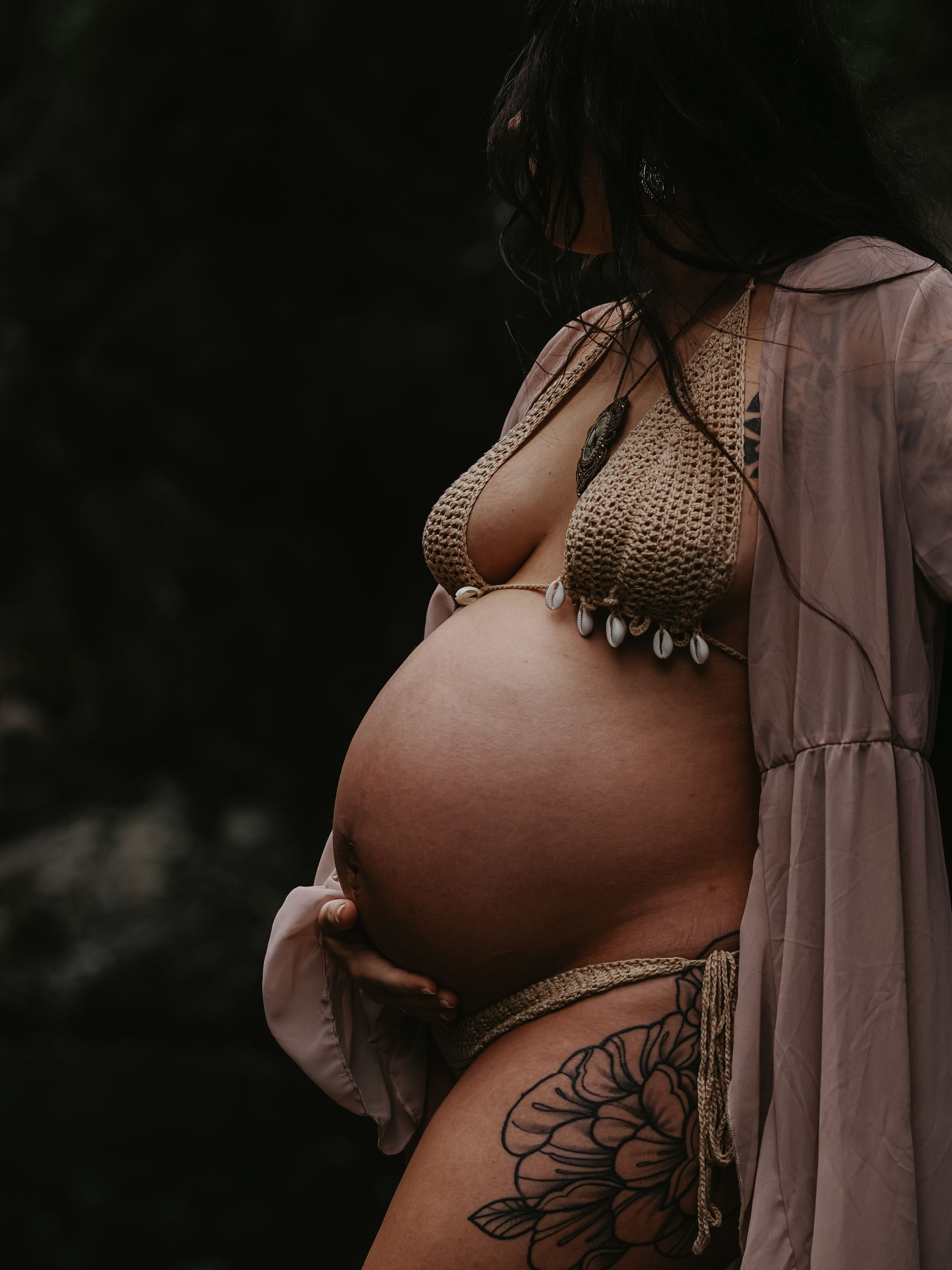 Pregnant person in a bikini, holding their belly. Tattoos on their leg and a light robe, dark background.