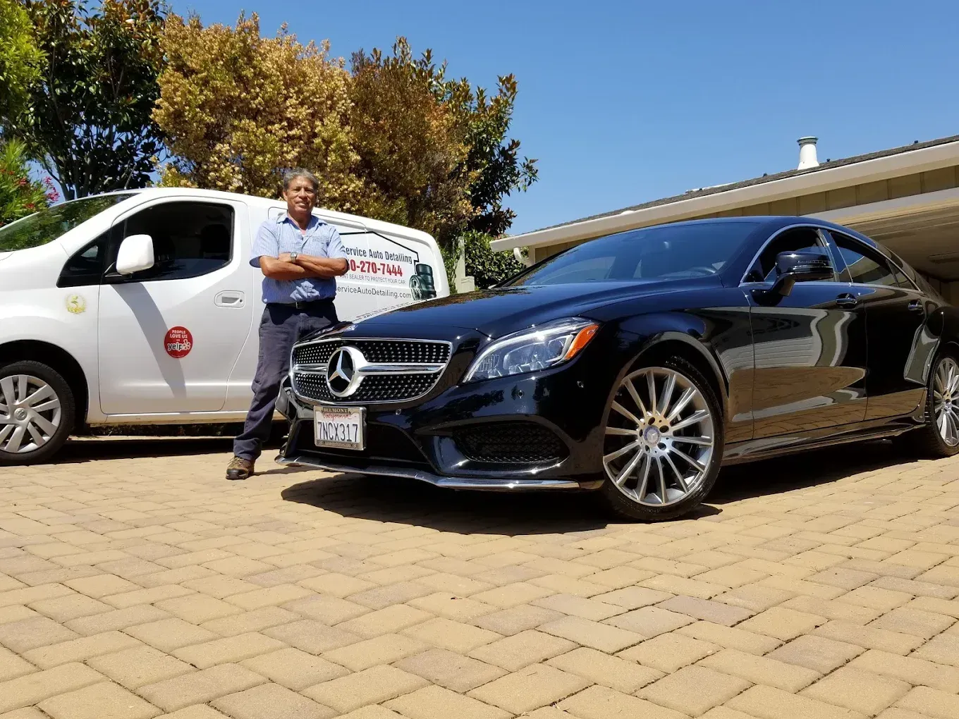 Man standing near a black Mercedes and a white van, on a brick driveway. Sunny day.