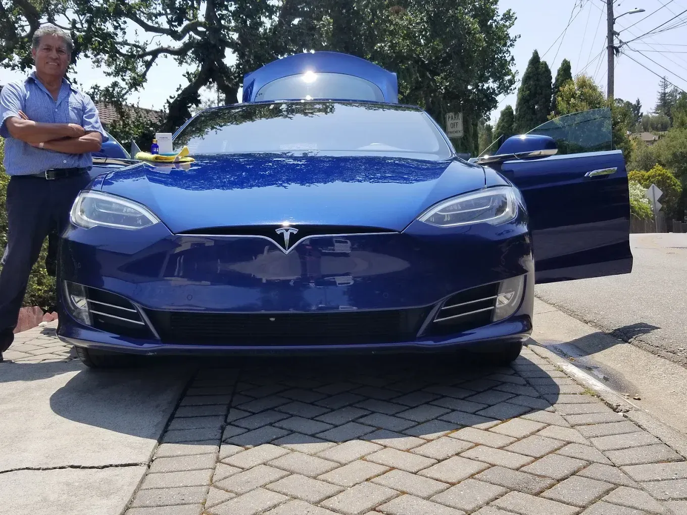 Man with arms crossed next to a blue Tesla with open doors on a sunny day.