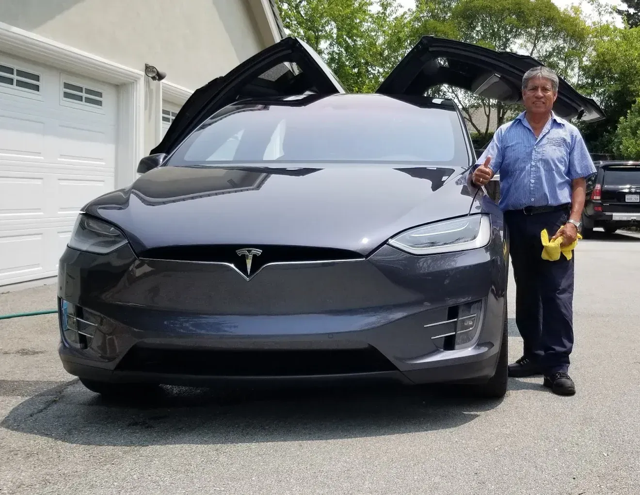 Man beside a gray Tesla Model X with falcon wing doors open, in front of a house.