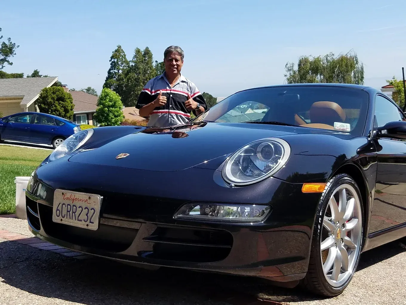 Man stands by a shiny black Porsche in a sunny residential setting.