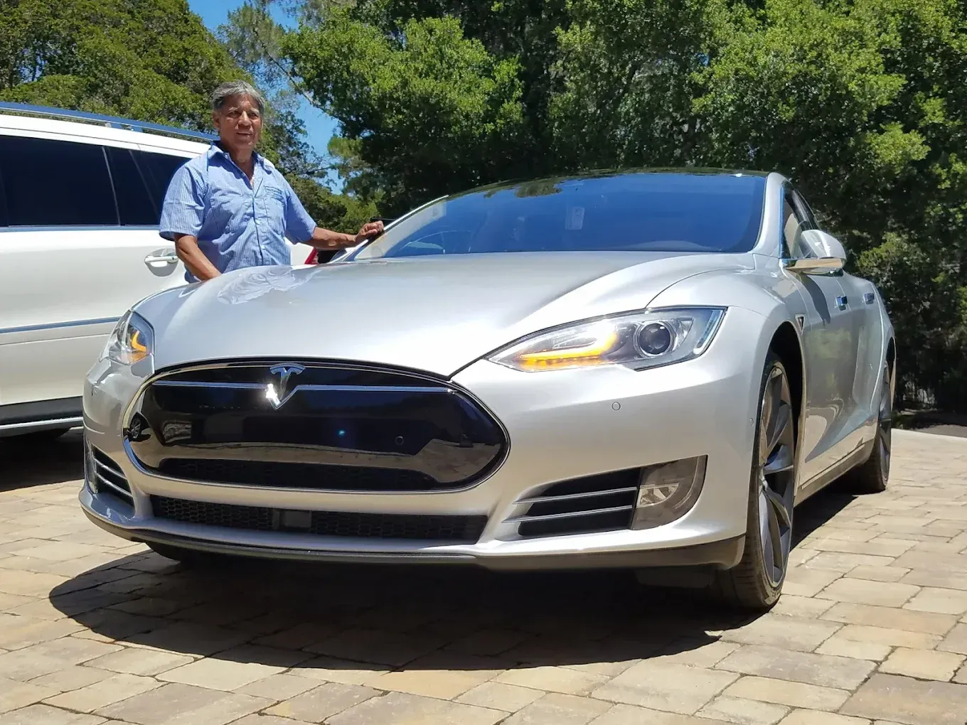 Man standing next to a silver Tesla sedan on a paved driveway; trees in the background.
