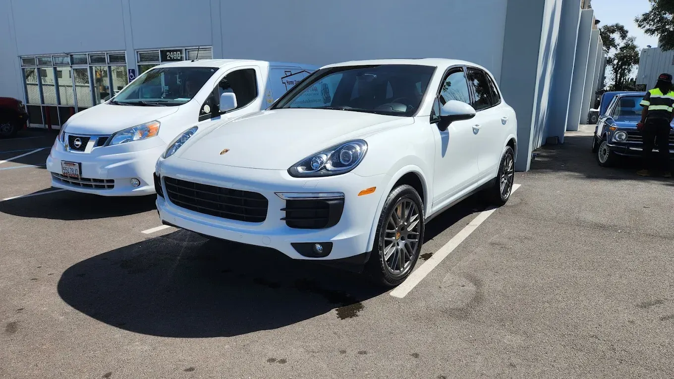 White Porsche SUV parked in a lot, beside a white van. Sunlight illuminates the vehicles.