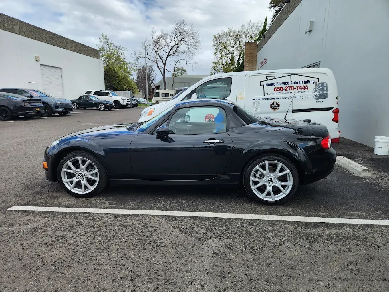 Dark blue Mazda Miata hardtop parked outdoors near a white van and buildings.