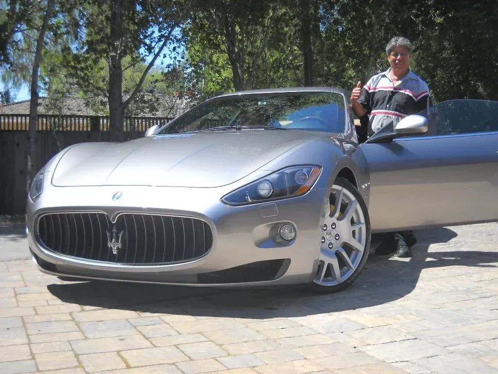 Man giving a thumbs up next to a silver Maserati convertible. Car is parked on a brick driveway.
