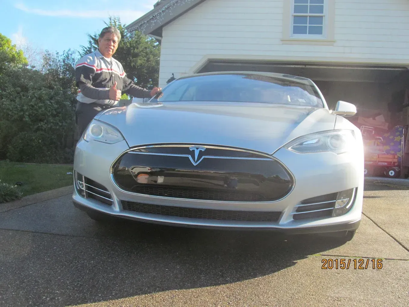 Man gives thumbs up next to a silver Tesla sedan parked in front of a garage.