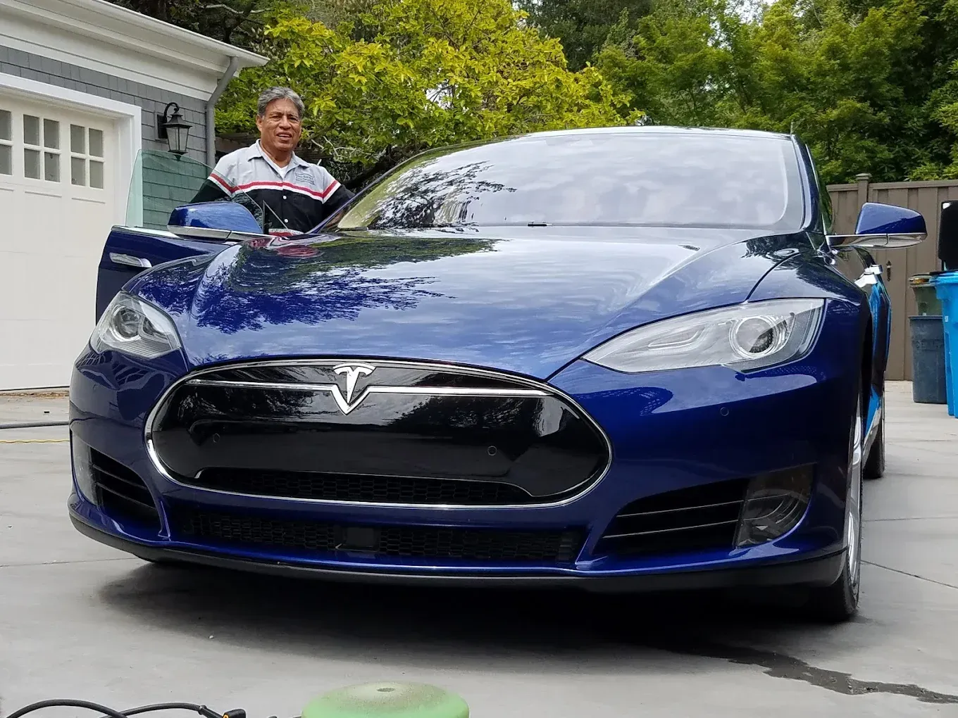Man washing a shiny blue Tesla sedan in a driveway.