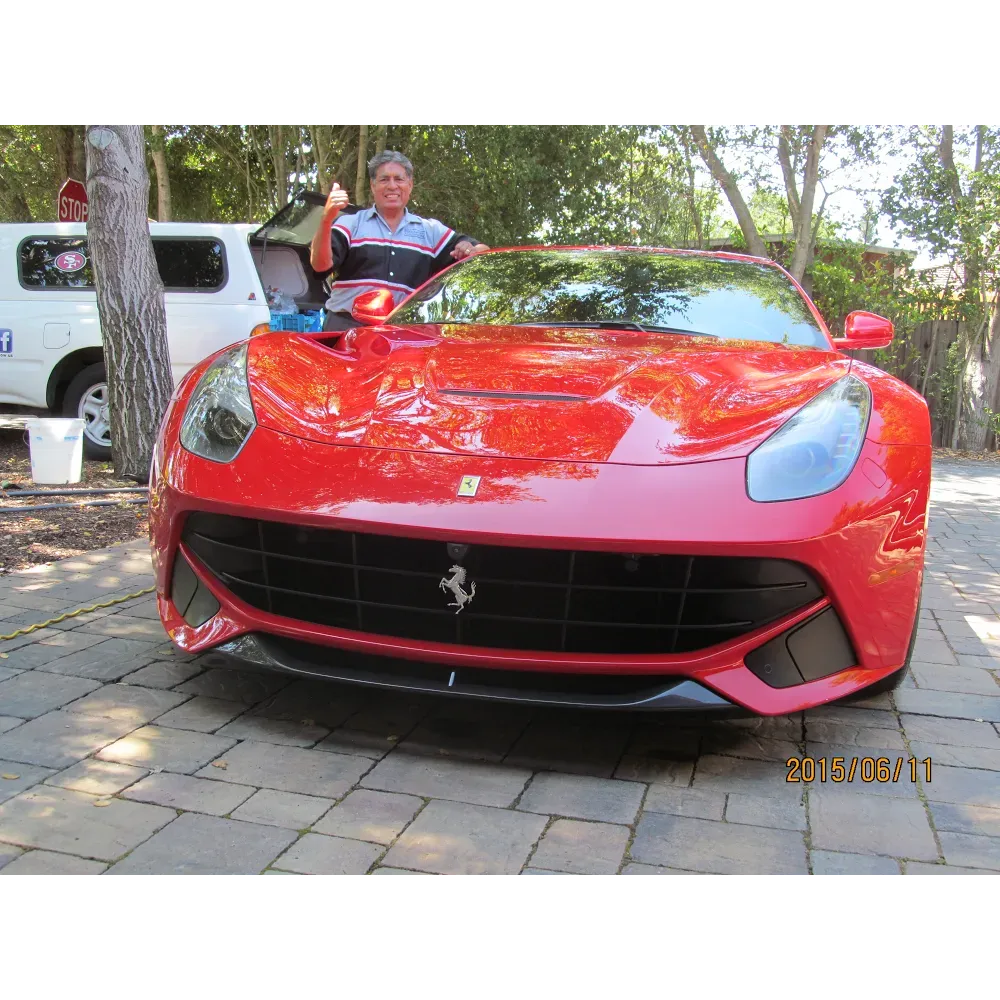 Man in red shirt gives thumbs up, stands beside red Ferrari. Paved driveway, trees in background.