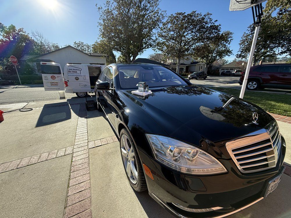 Black Mercedes-Benz sedan parked in front of a house on a sunny day. A service van is nearby.