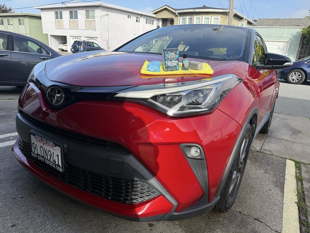 Red Toyota C-HR with items on hood; parked on street in front of apartment buildings.