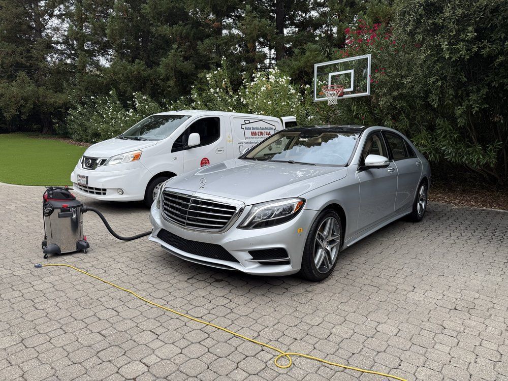 A silver sedan car being detailed by a vacuum connected to a white service van in a driveway.