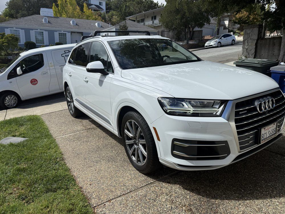 White Audi SUV parked on a driveway next to a white van, houses in the background.