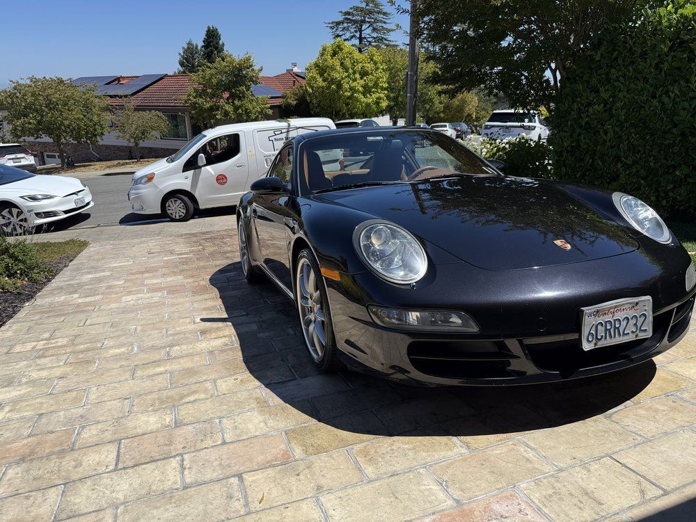 Black Porsche 911 parked on a brick driveway. A white van is in the background.