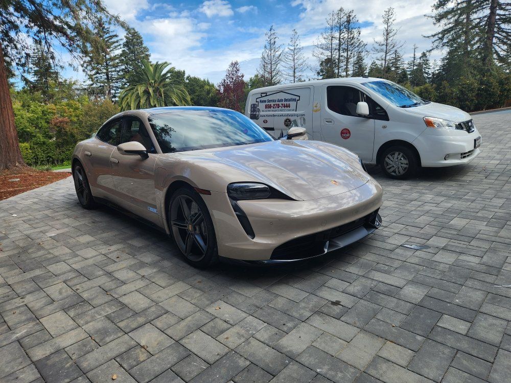 Tan Porsche electric car parked next to a white service van on a paved driveway with trees in the background.