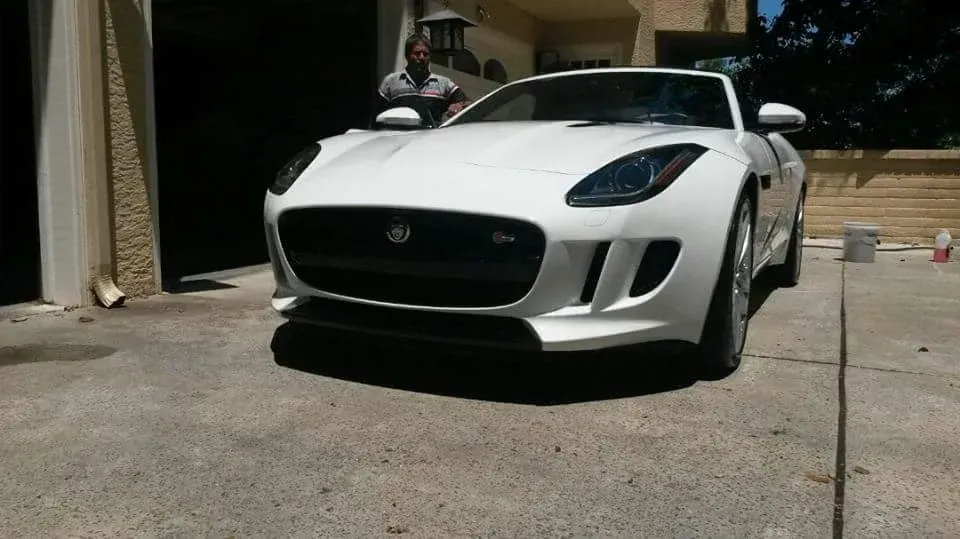 White Jaguar F-Type parked in a driveway. A person stands nearby. Sunny day.