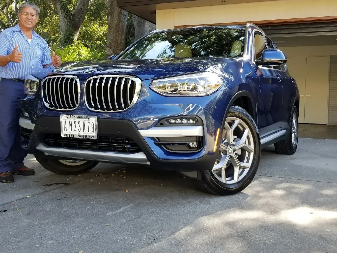 Man giving thumbs up next to a blue BMW SUV parked on a driveway.