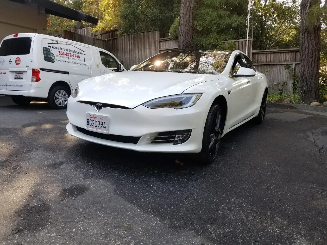 White Tesla sedan parked in front of a white service van on an asphalt driveway with trees in the background.