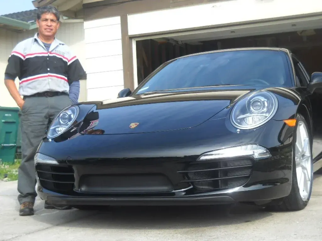 Man in mechanic shirt stands next to a black Porsche in front of a garage.