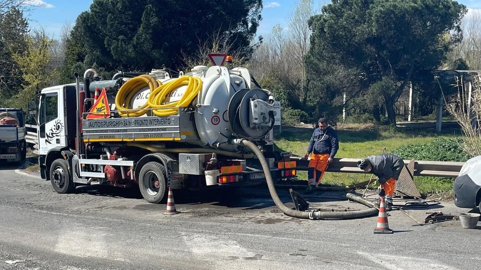 Un camion bianco per la pulizia delle fognature è parcheggiato a bordo strada, con due operai e l'attrezzatura necessaria per pulire un'area di drenaggio.
