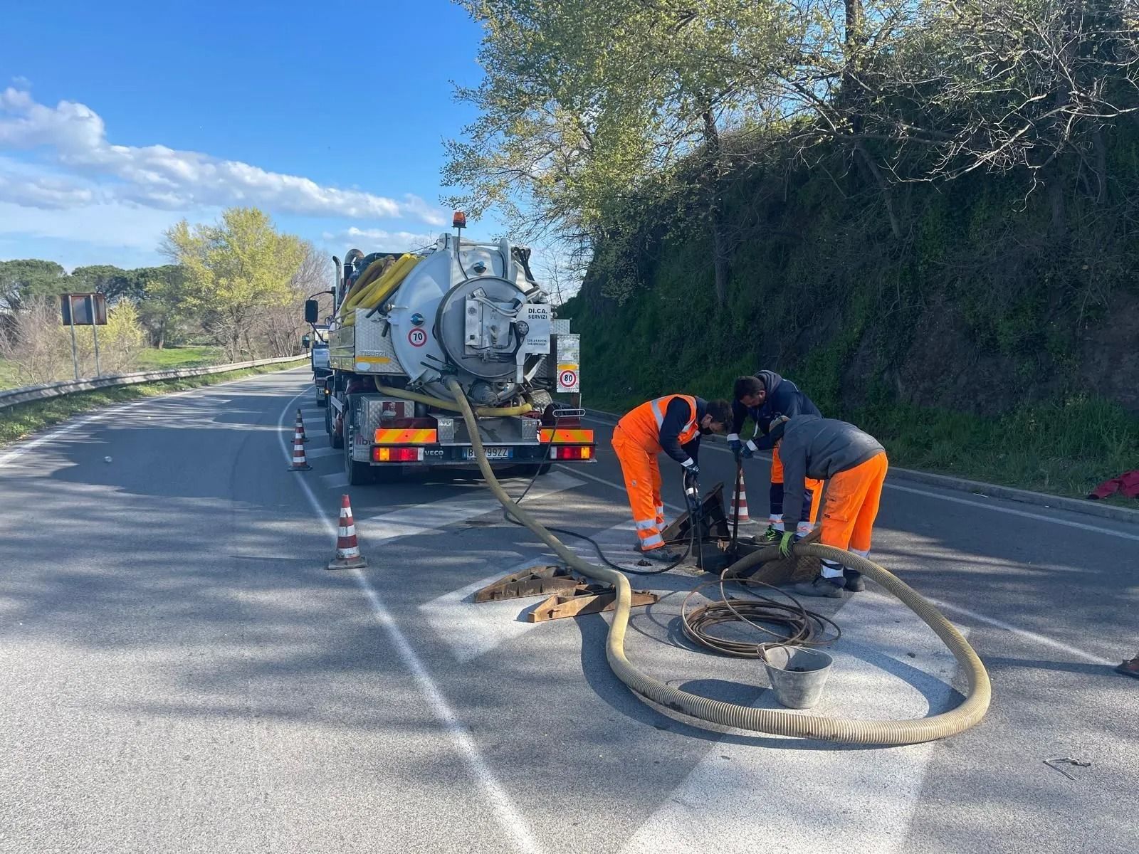 Alcuni operai con indumenti di sicurezza arancioni utilizzano un camion aspiratore per pulire una griglia di scolo sul ciglio di una soleggiata strada rurale.