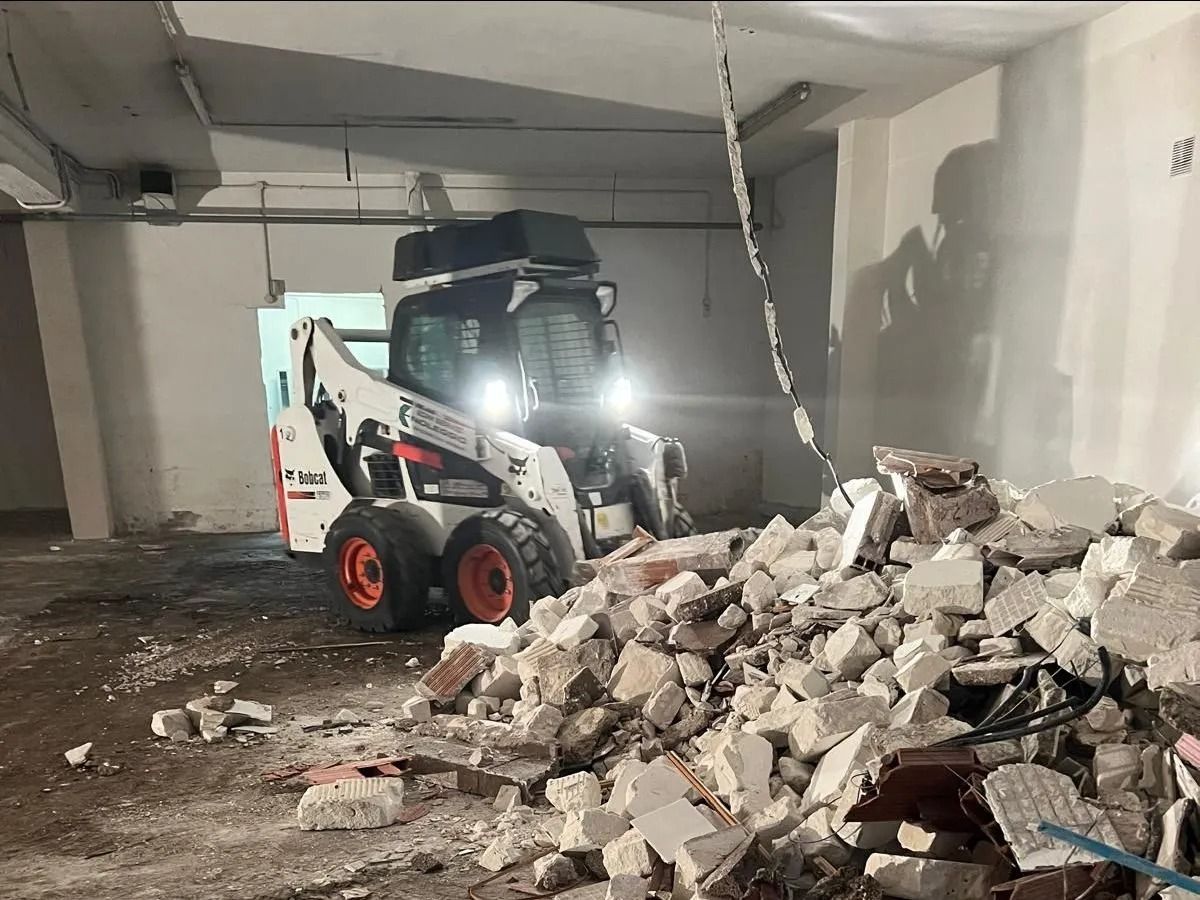 Una pala caricatrice compatta bianca rimuove un cumulo di detriti edili all'interno di una stanza in cemento grezzo.