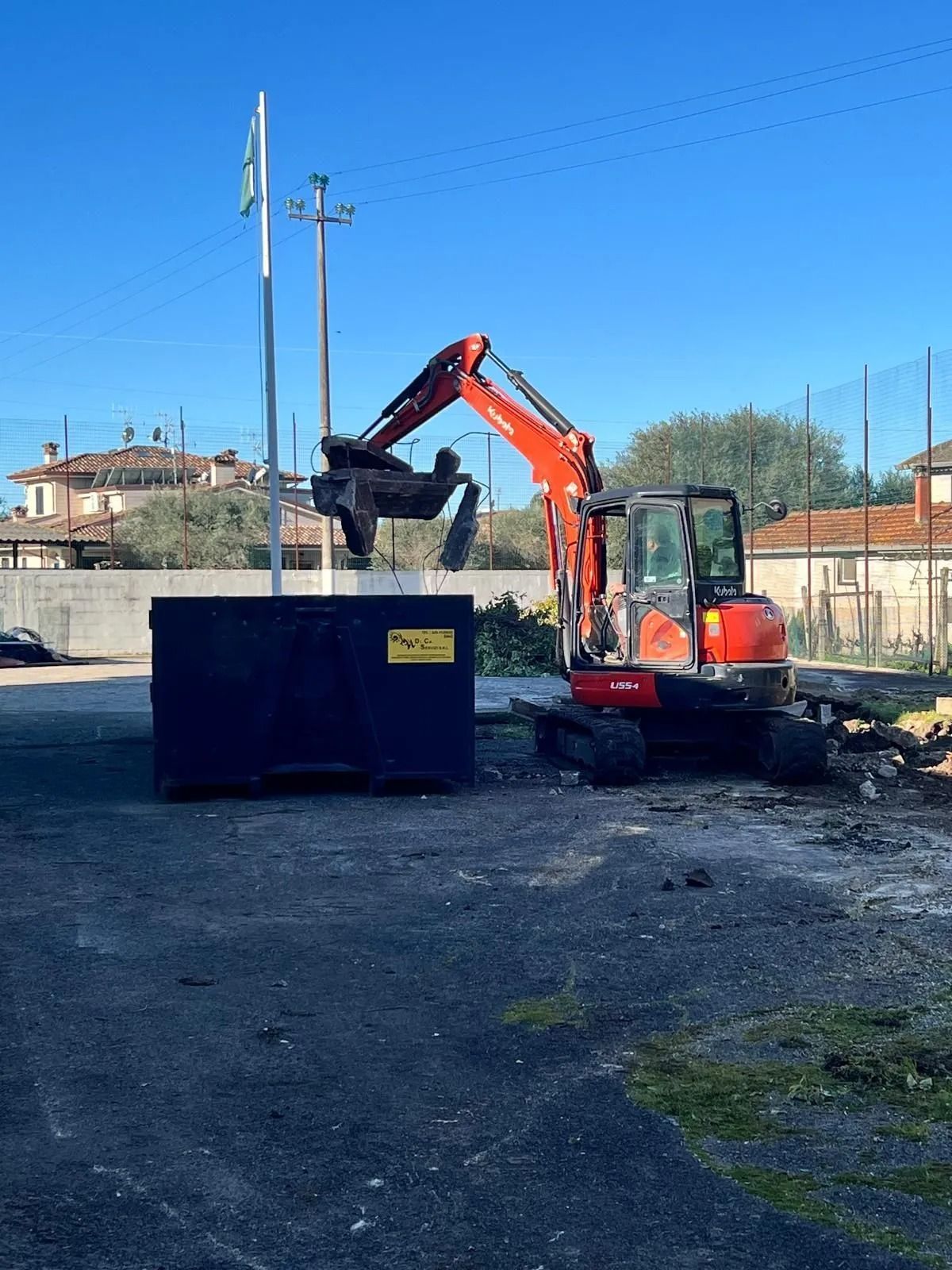 Un escavatore rosso solleva un oggetto pesante e lo deposita in un grande cassonetto blu su un piazzale ghiaioso, sotto un cielo azzurro e limpido.