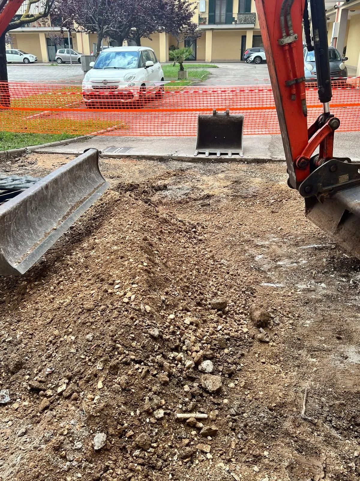 Un escavatore arancione brillante con una benna staccata parcheggiato in un cantiere sterrato, con delle auto sullo sfondo.