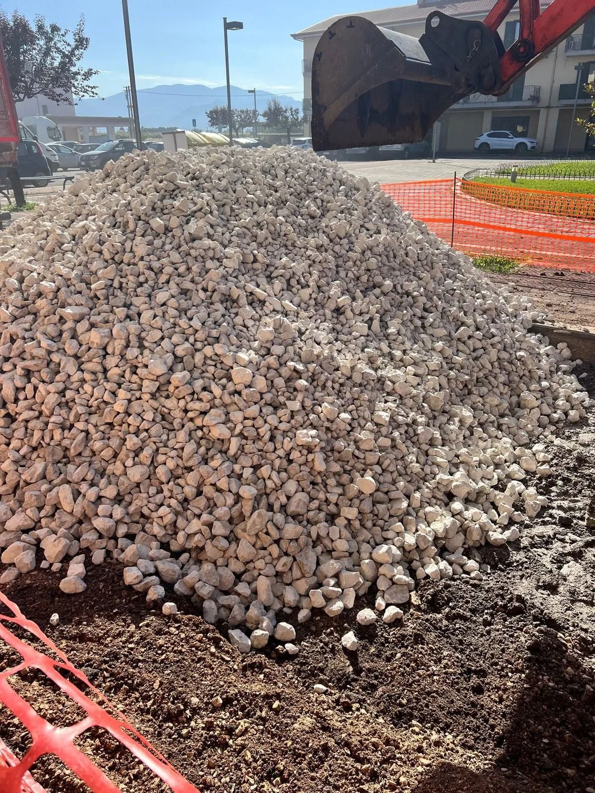 In un cantiere edile, un grande cumulo di ghiaia chiara si erge su un terreno scuro, con la benna di un escavatore ben visibile sopra di esso.