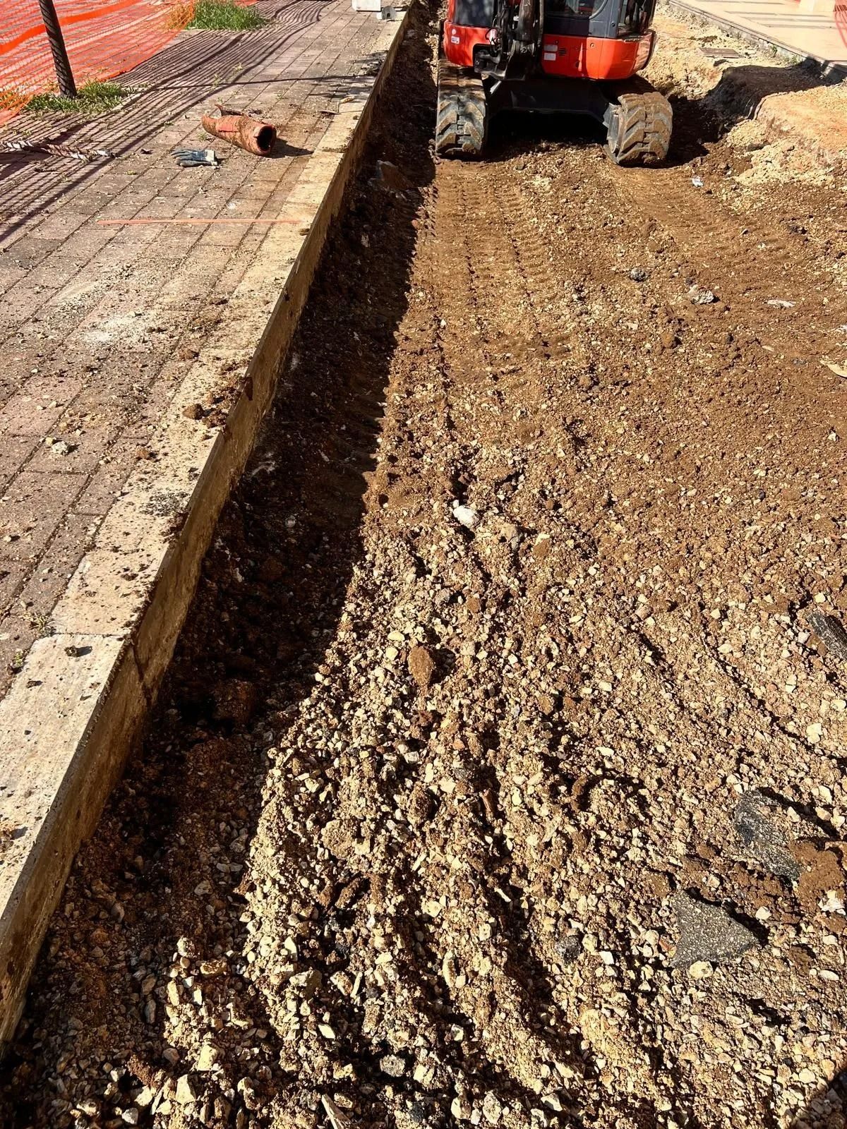 In un cantiere edile si vede uno scavo in corso accanto a un marciapiede, con un piccolo escavatore arancione visibile sullo sfondo.
