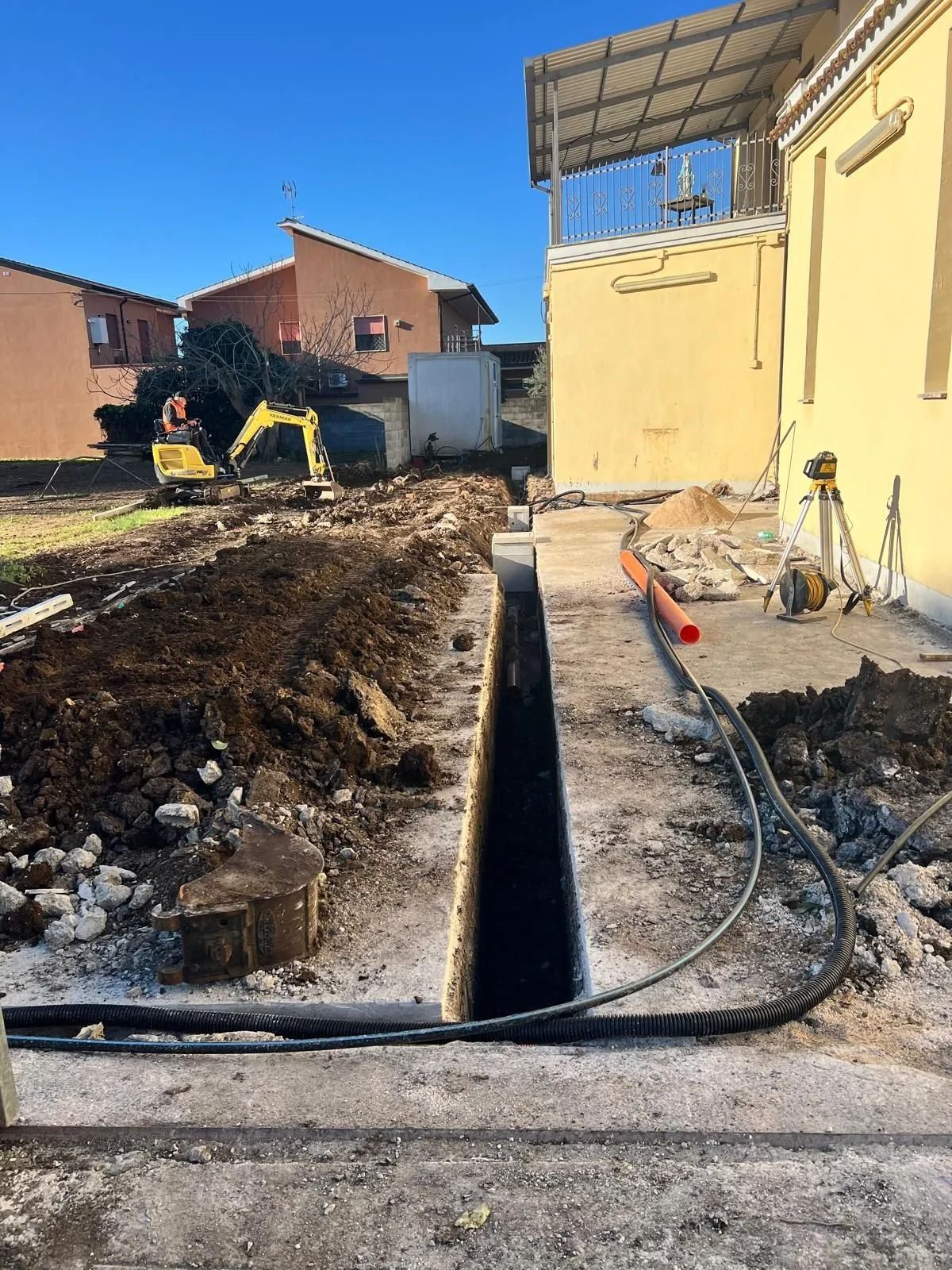 Una stretta trincea scavata di fronte a un edificio giallo, con un piccolo escavatore giallo sullo sfondo.