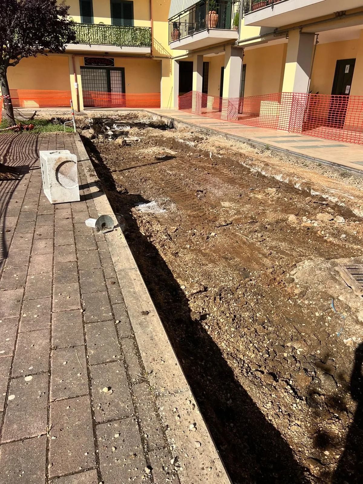 Un cantiere edile di fronte a un edificio a più piani, caratterizzato da uno scavo di terra battuta delimitato da lastre di pietra.