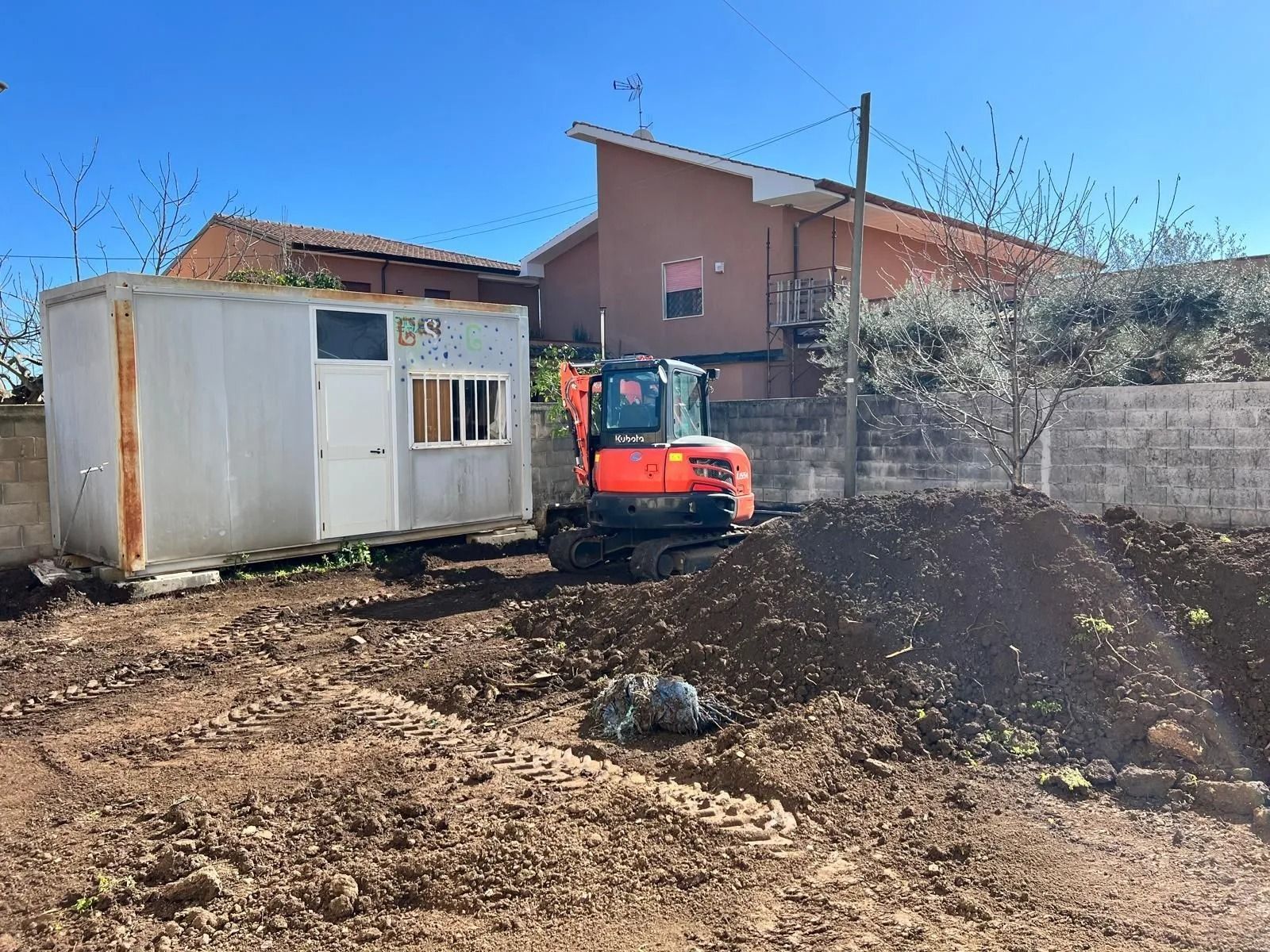 Un miniescavatore arancione è posizionato su un terreno sterrato accanto a un capannone rettangolare bianco, con una casa visibile sullo sfondo.
