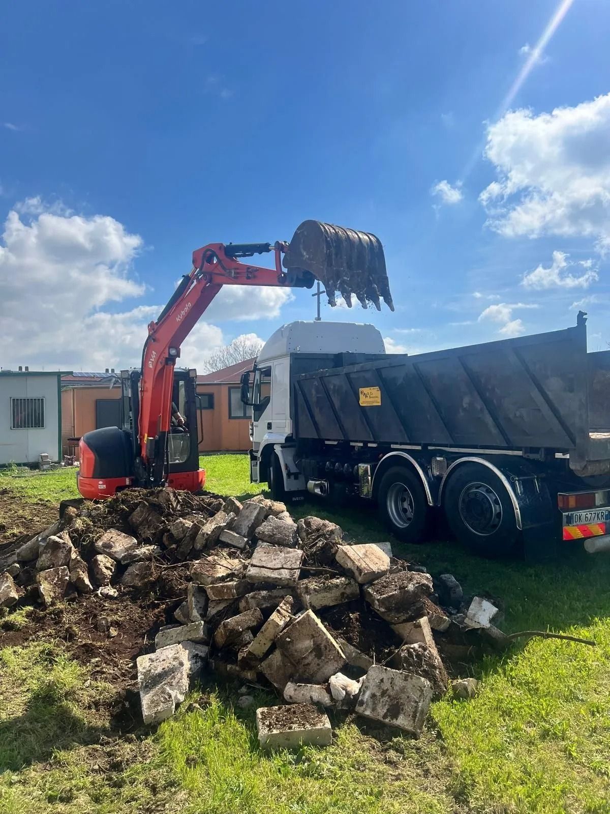 In una giornata di sole, un escavatore arancione riempie il cassone di un autocarro bianco con cemento frantumato.