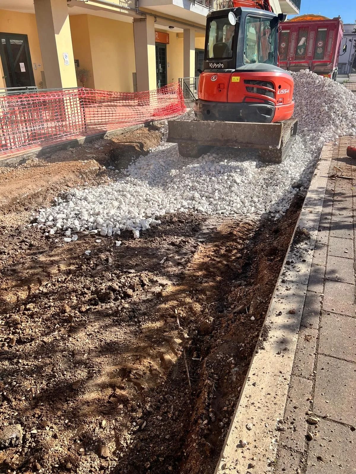 In pieno giorno, un escavatore rosso livella la ghiaia in un cantiere edile accanto a un marciapiede e a un edificio.