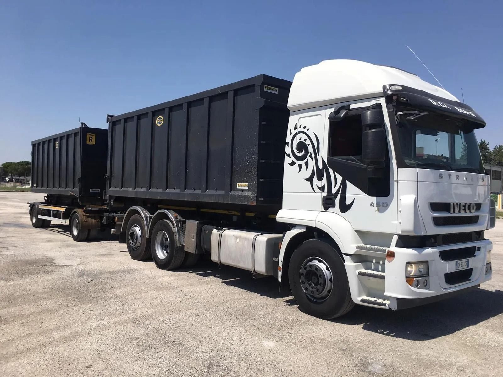 Un camion Iveco bianco con rimorchio abbinato e due container neri parcheggiati su un piazzale ghiaioso sotto un cielo azzurro e limpido.