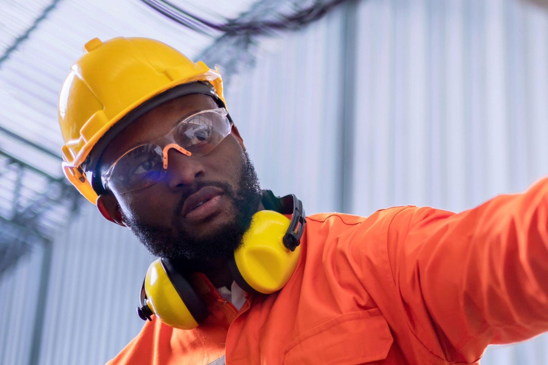 Un hombre con ropa de trabajo naranja, casco y protección para los oídos mira hacia arriba, usando gafas de seguridad en el interior.