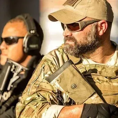 Dos hombres armados, uno con gafas de sol y gorra color canela, con aspecto alerta. El otro lleva auriculares y gafas de sol.