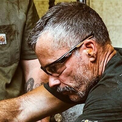 Hombre con cabello canoso y bigote, usando gafas de seguridad y tapones para los oídos, trabajando.