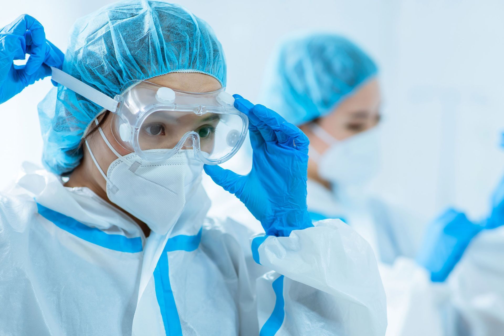 Persona con traje protector ajustándose las gafas protectoras en un laboratorio. Otra persona con atuendo similar al fondo.