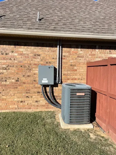 An air conditioning unit next to a brick wall with an electrical box, and a brown fence.