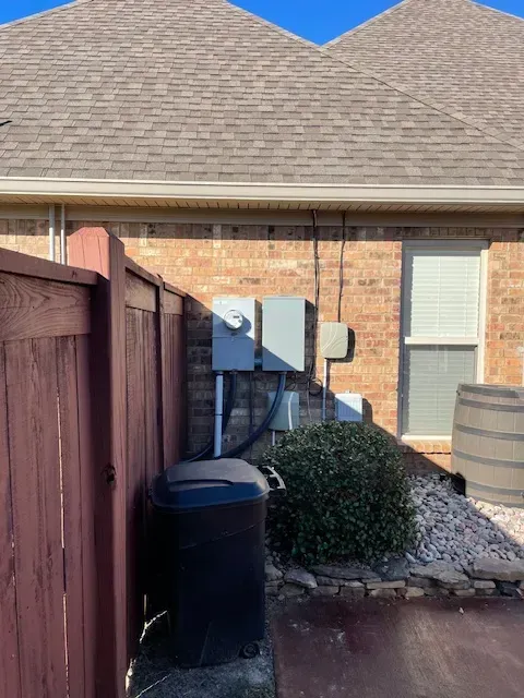 Electrical boxes mounted on a brick wall, next to a window. A dark trash can sits in front of the wall.
