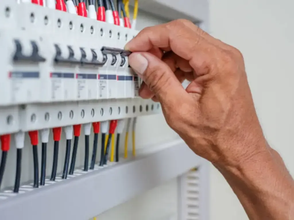 Hand flipping a circuit breaker switch in a gray electrical panel with red and black wires.