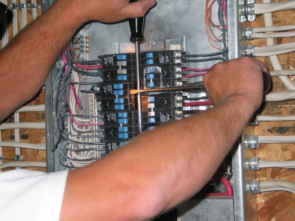 Person working on an electrical panel with a screwdriver. Wires and circuit breakers are visible.