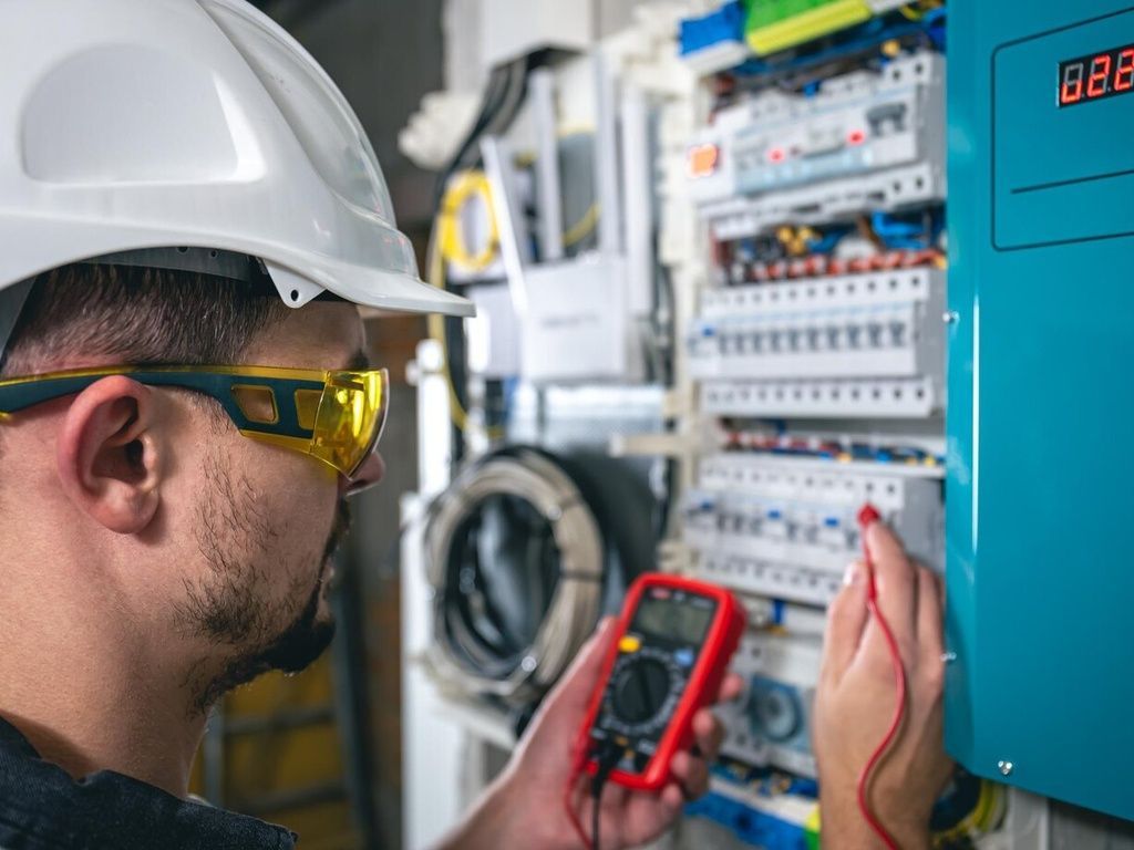 Electrician in safety gear tests a circuit panel with a multimeter.