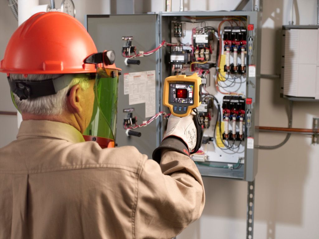 An electrician using a thermal imager on an electrical panel, wearing safety gear.