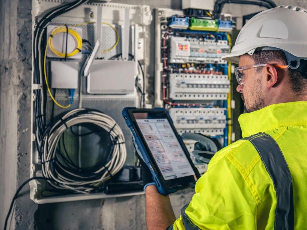 Electrician in a safety vest and hardhat, using a tablet to inspect wiring in an electrical panel.