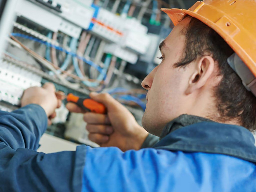 Electrician in an orange hardhat working on electrical panel with screwdriver.