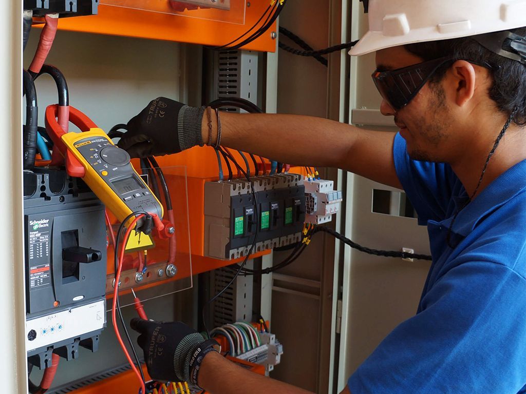 Electrician in safety gear tests wires in an orange electrical panel with a multimeter.