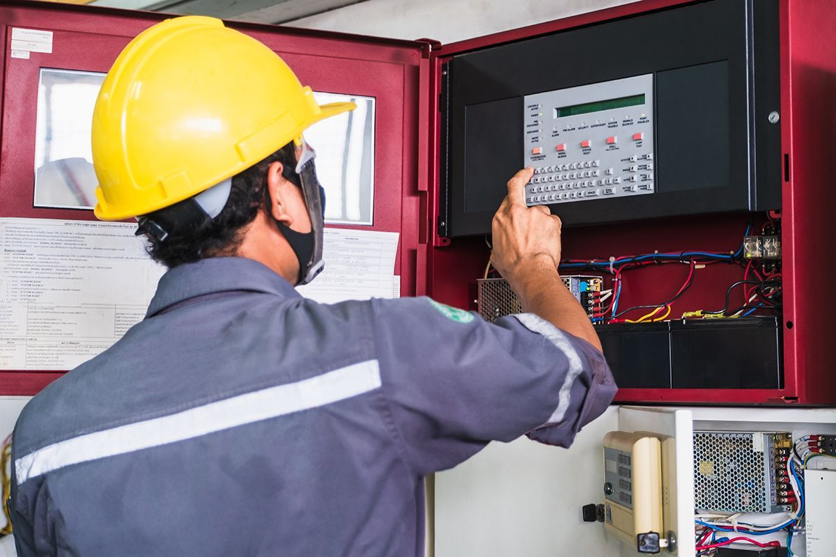 Worker in a yellow hard hat and mask, operating a fire alarm panel inside a red cabinet.